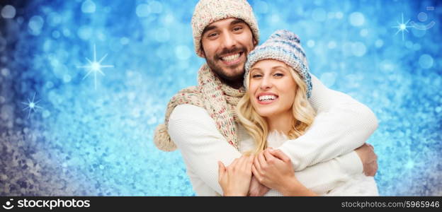 winter, fashion, couple, christmas and people concept - smiling man and woman in hats and scarf hugging over blue holidays lights or glitter background