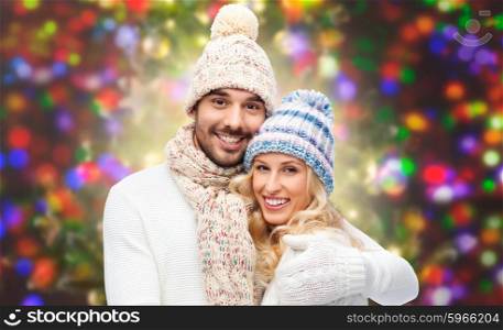 winter, fashion, couple, christmas and people concept - smiling man and woman in hats and scarf hugging over holidays lights background