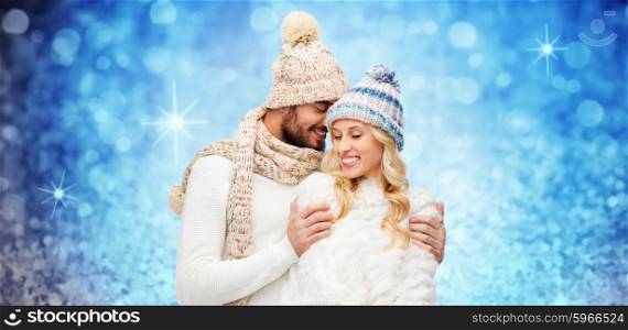 winter, fashion, couple, christmas and people concept - smiling man and woman in hats and scarf hugging over blue holidays lights or glitter background