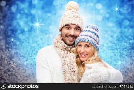 winter, fashion, couple, christmas and people concept - smiling man and woman in hats and scarf hugging over blue glitter and holidays lights background