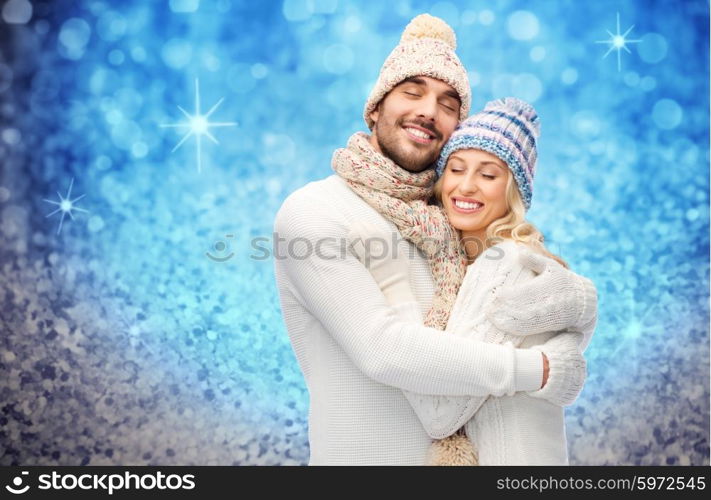 winter, fashion, couple, christmas and people concept - smiling man and woman in hats and scarf hugging over blue glitter or lights background