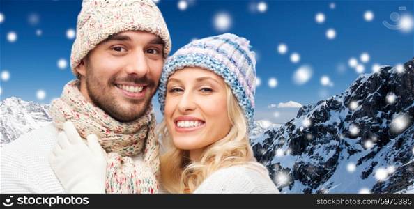 winter, fashion, couple, christmas and people concept - smiling man and woman in hats and scarf hugging over snow and mountains background