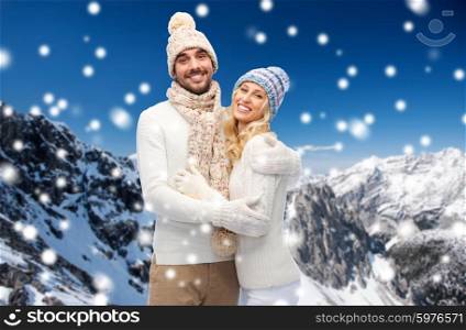 winter, fashion, couple, christmas and people concept - smiling man and woman in hats and scarf hugging over snowy mountains background