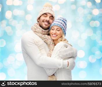winter, fashion, couple, christmas and people concept - smiling man and woman in hats and scarf hugging over blue holidays lights background