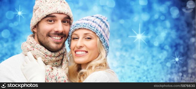 winter, fashion, couple, christmas and people concept - smiling man and woman in hats and scarf hugging over blue glitter or holidays lights background