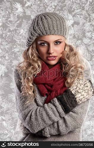 winter fashion shot of a beautiful girl with long curled blonde hair wearing a grey woolen cap, a grey sweater and warm gloves