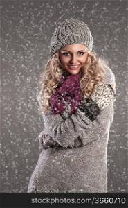 winter fashion shot of a smiling attractive blonde wearing a wool cap, a grey wool sweater, gloves and a purple scarf