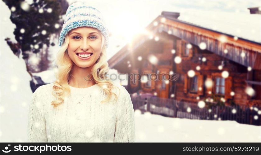 winter, fashion, vacation, christmas and people concept - smiling young woman in hat and sweater over wooden country house and snowflakes background. smiling young woman in winter hat and sweater