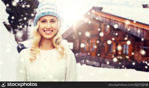 winter, fashion, vacation, christmas and people concept - smiling young woman in hat and sweater over wooden country house and snowflakes background. smiling young woman in winter hat and sweater