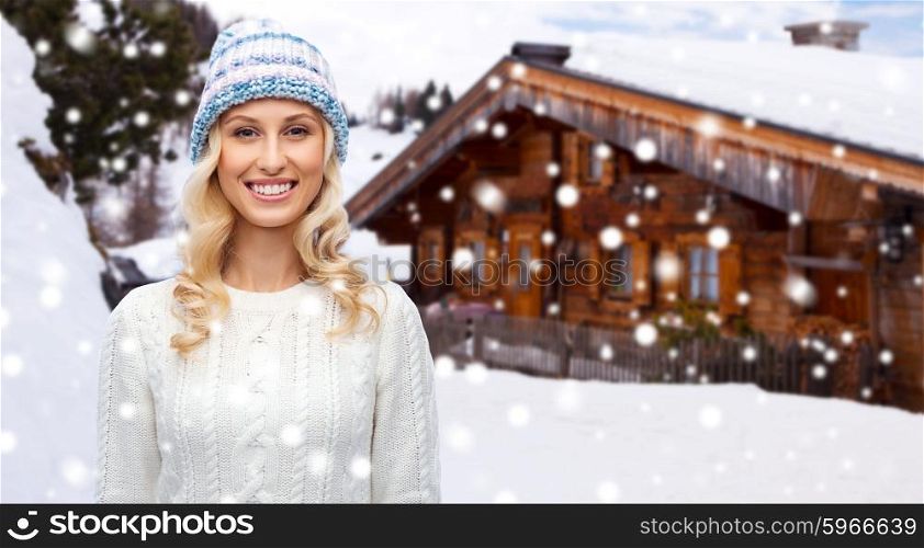 winter, fashion, vacation, christmas and people concept - smiling young woman in hat and sweater over wooden country house and snowflakes background