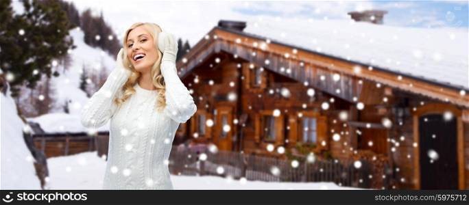 winter, fashion, vacation, christmas and people concept - smiling young woman in earmuffs and sweater over wooden country house and snow background