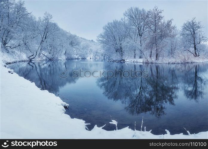 Winter forest on the river at sunset. Colorful landscape with snowy trees, river with reflection in water in cold evening. Snow covered trees, lake, sun and blue sky. Beautiful forest in snowy winter. Landscape with snowy trees in winter forest