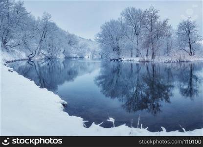 Winter forest on the river at sunset. Colorful landscape with snowy trees, river with reflection in water in cold evening. Snow covered trees, lake, sun and blue sky. Beautiful forest in snowy winter. Landscape with snowy trees in winter forest