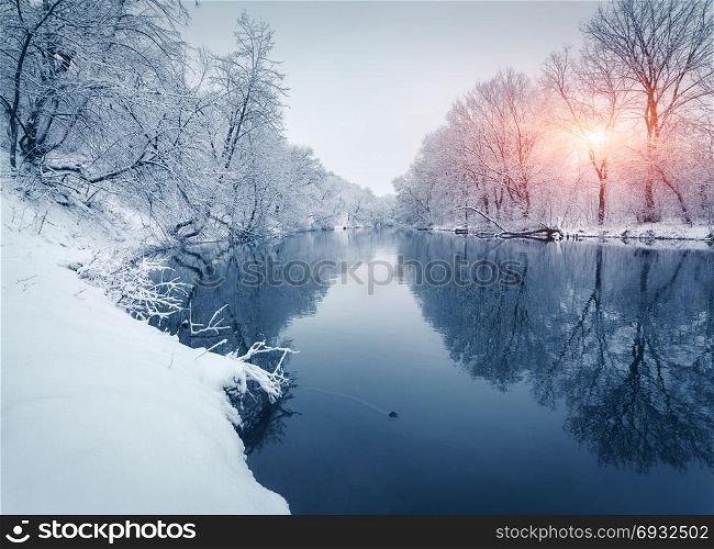 Winter forest on the river at sunset. Colorful landscape with snowy trees, frozen river with reflection in water. Seasonal. Snow covered trees, lake, sun and blue sky. Beautiful forest in snowy winter. Winter forest on the river at sunset. Colorful landscape with snowy trees