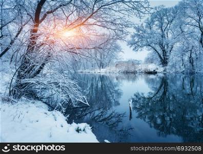 Winter forest on the river at sunset. Colorful landscape with snowy trees, frozen river with reflection in water. Seasonal. Snow covered trees, lake, sun and blue sky. Beautiful forest in snowy winter. Winter forest on the river at sunset. Colorful landscape with snowy trees