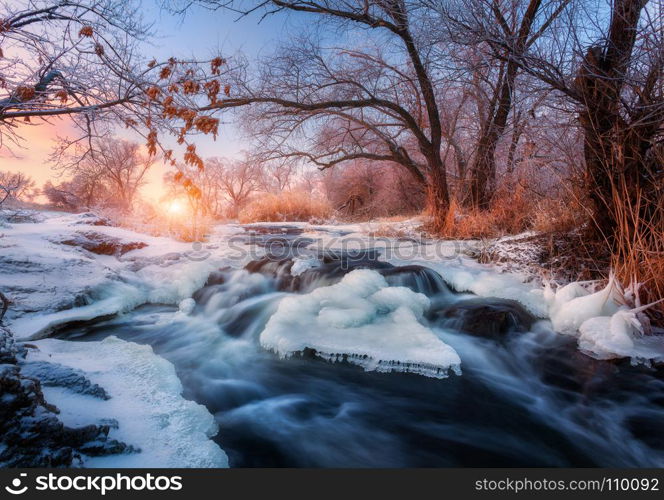 Winter forest with amazing river at sunset. Winter landscape with snowy trees, ice, beautiful frozen river, snowy bushes, colorful sky in dusk. Blurred water. Small waterfalls.Creek with water cascade. Winter landscape with snowy trees, ice, beautiful frozen river