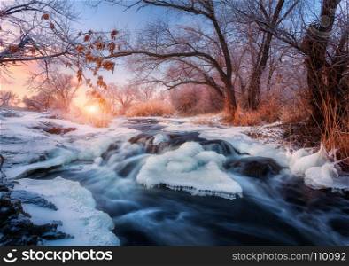 Winter forest with amazing river at sunset. Winter landscape with snowy trees, ice, beautiful frozen river, snowy bushes, colorful sky in dusk. Blurred water. Small waterfalls.Creek with water cascade. Winter landscape with snowy trees, ice, beautiful frozen river