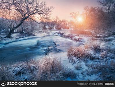 Winter forest with amazing river at sunset. Winter landscape with snowy trees, ice, beautiful frozen river, snowy bushes, colorful sky in dusk. Blurred water. Small waterfalls.Creek with water cascade. Winter landscape with snowy trees, ice, beautiful frozen river