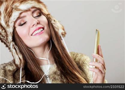 Winter girl listening music using phone with headphones. Happy woman wearing fur vest and warm hat in freezing cold time. . Winter girl enjoying music.