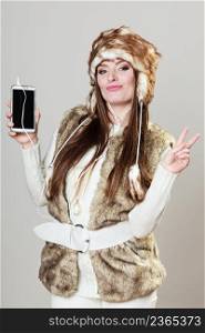 Winter girl listening music using phone with headphones giving peace gesture. Happy woman wearing fur vest and warm hat in freezing cold time. . Winter girl listening music.