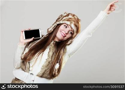 Winter girl listening music using phone with headphones raising hand. Happy woman wearing fur vest and warm hat in freezing cold time having fun posing. . Winter girl listening music.