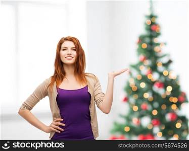 winter holidays, advertising and people concept - smiling teenage girl in casual clothes holding something on her palm over living room and christmas tree background