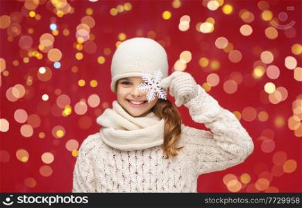 winter holidays and celebration concept - girl in hat, muffler and gloves with big snowflake over christmas lights on red background. girl with big snowflake over christmas lights