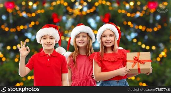 winter holidays and childhood concept - happy smiling children in santa helper hats showing ok hand sign with gift box over christmas tree lights background. happy smiling children with christmas gift
