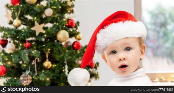 winter holidays and childhood concept - lovely baby boy in santa hat over christmas tree at home. lovely baby boy over christmas tree at home