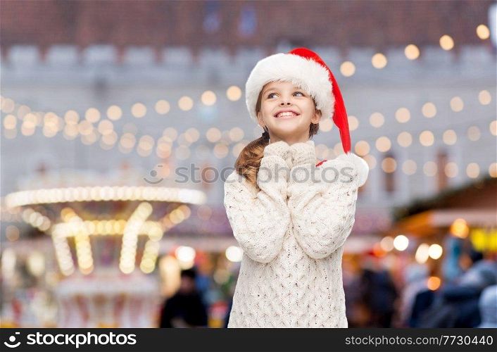 winter holidays and children concept - dreaming girl in santa helper hat over christmas market or amusement park background. happy smiling girl dreaming over christmas market