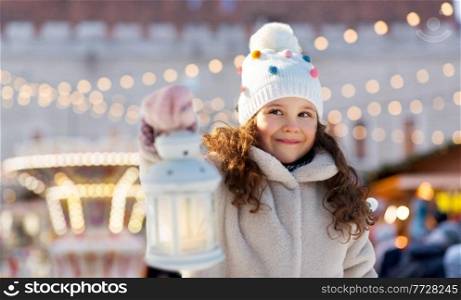 winter holidays and children concept - portrait of happy little girl with lantern over christmas market or amusement park background. happy little girl with christmas lantern in winter
