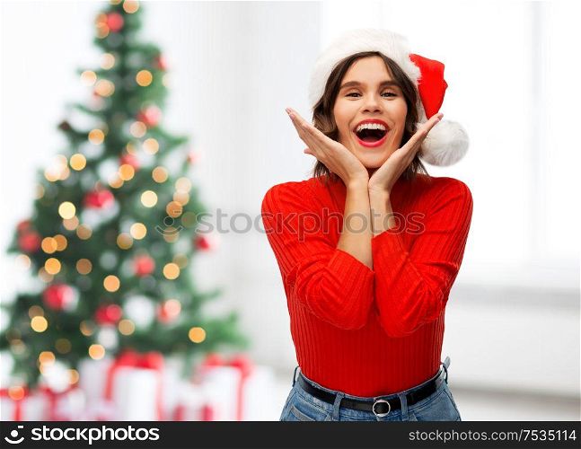 winter holidays and emotions concept - happy smiling young woman in santa helper hat over christmas tree lights background. happy young woman in santa hat on christmas