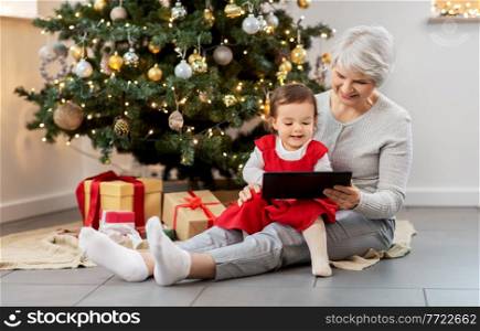 winter holidays and family concept - happy grandmother and baby granddaughter with tablet pc computer sitting near christmas tree at home. grandmother and baby girl with christmas gifts
