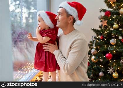winter holidays and family concept - happy middle-aged father and baby daughter looking through window over christmas tree at home. father and baby daughter on christmas at home