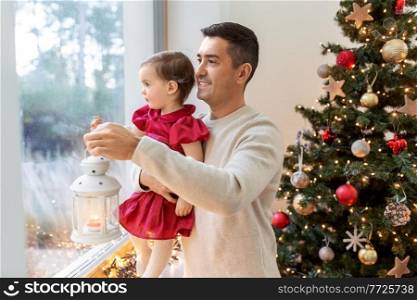 winter holidays and family concept - happy middle-aged father with lantern and baby daughter looking through window over christmas tree at home. father and baby daughter on christmas at home
