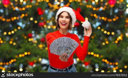 winter holidays and finance concept - happy smiling young woman in santa helper hat holding dollar money banknotes over christmas tree lights on background. happy woman in santa hat with money on christmas