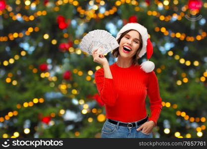 winter holidays and finance concept - happy smiling young woman in santa helper hat holding dollar money banknotes over christmas tree lights background. happy woman in santa hat with money on christmas