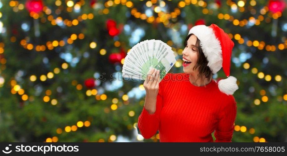 winter holidays and finance concept - happy smiling young woman in santa helper hat holding euro money banknotes over christmas tree lights on background. happy woman in santa hat with money on christmas