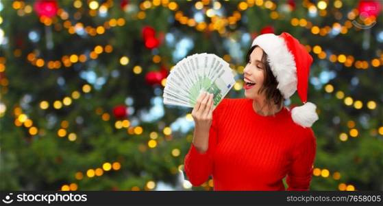 winter holidays and finance concept - happy smiling young woman in santa helper hat holding euro money banknotes over christmas tree lights on background. happy woman in santa hat with money on christmas