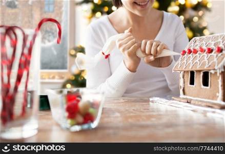 winter holidays and food cooking concept - close up of happy smiling woman with baking bag making gingerbread house at home over christmas tree lights background. close up of woman making gingerbread house