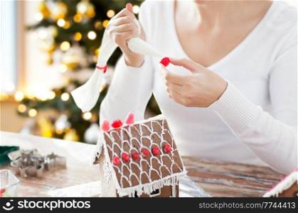 winter holidays and food cooking concept - close up of woman with baking bag making gingerbread house at home over christmas tree lights background. close up of woman making gingerbread house
