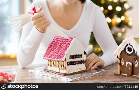 winter holidays and food cooking concept - close up of woman with baking bag making gingerbread house at home over christmas tree lights background. close up of woman making gingerbread house