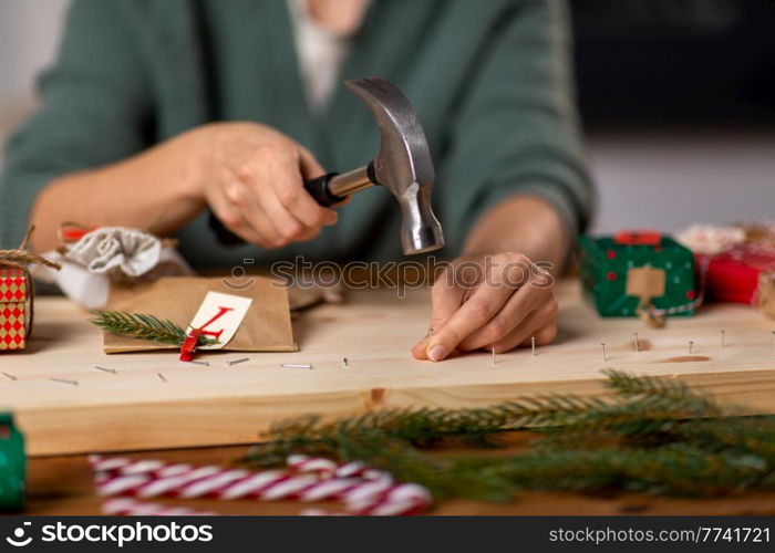 winter holidays and hobby concept - close up of woman with hammer hammering nails in wooden board and making advent calendar at home. woman making advent calendar on christmas at home