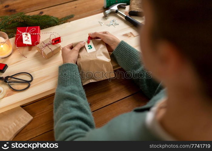 winter holidays and hobby concept - hands with clothespin, craft paper bag and tag making advent calendar on christmas at home. hands making christmas advent calendar at home