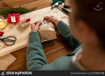 winter holidays and hobby concept - hands with clothespin, craft paper bag and tag making advent calendar on christmas at home. hands making christmas advent calendar at home