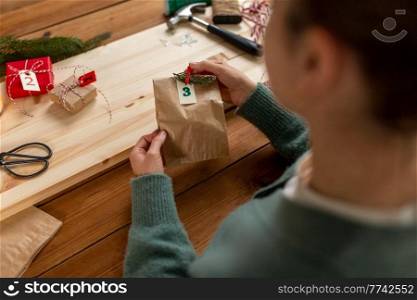 winter holidays and hobby concept - hands with clothespin, craft paper bag and tag making advent calendar on christmas at home. hands making christmas advent calendar at home