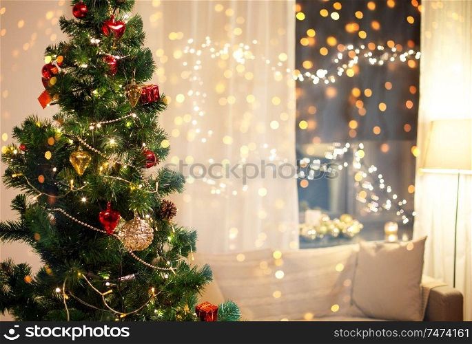 winter holidays and interior concept - close up of decorated christmas tree, sofa and garland on window at home in evening. close up of christmas tree at home in evening