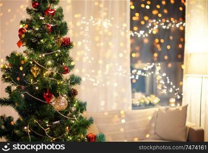 winter holidays and interior concept - close up of decorated christmas tree, sofa and garland on window at home in evening. close up of christmas tree at home in evening