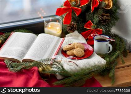 winter holidays and leisure concept - open book, fir branch, oatmeal cookies, cup of coffee and christmas decorations on window sill at home. book, cookies, glasses, coffee and christmas decor