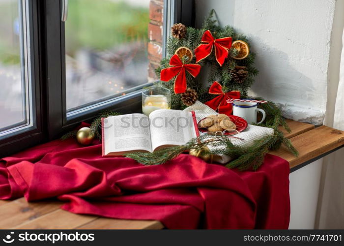 winter holidays and leisure concept - open book, fir branch, oatmeal cookies, coffee and christmas decorations on window sill at home. book, cookies, cup of coffee and christmas decor